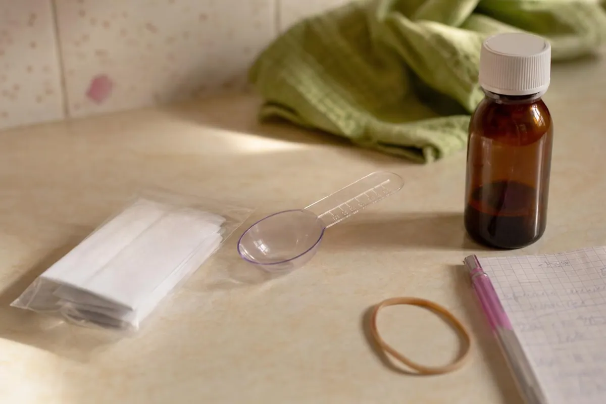 Gros plan sur une pochette plastique contenant une ordonnance pliee, une cuillere doseuse et une feuille d’autorisation manuscrite posees sur un plan de travail clair